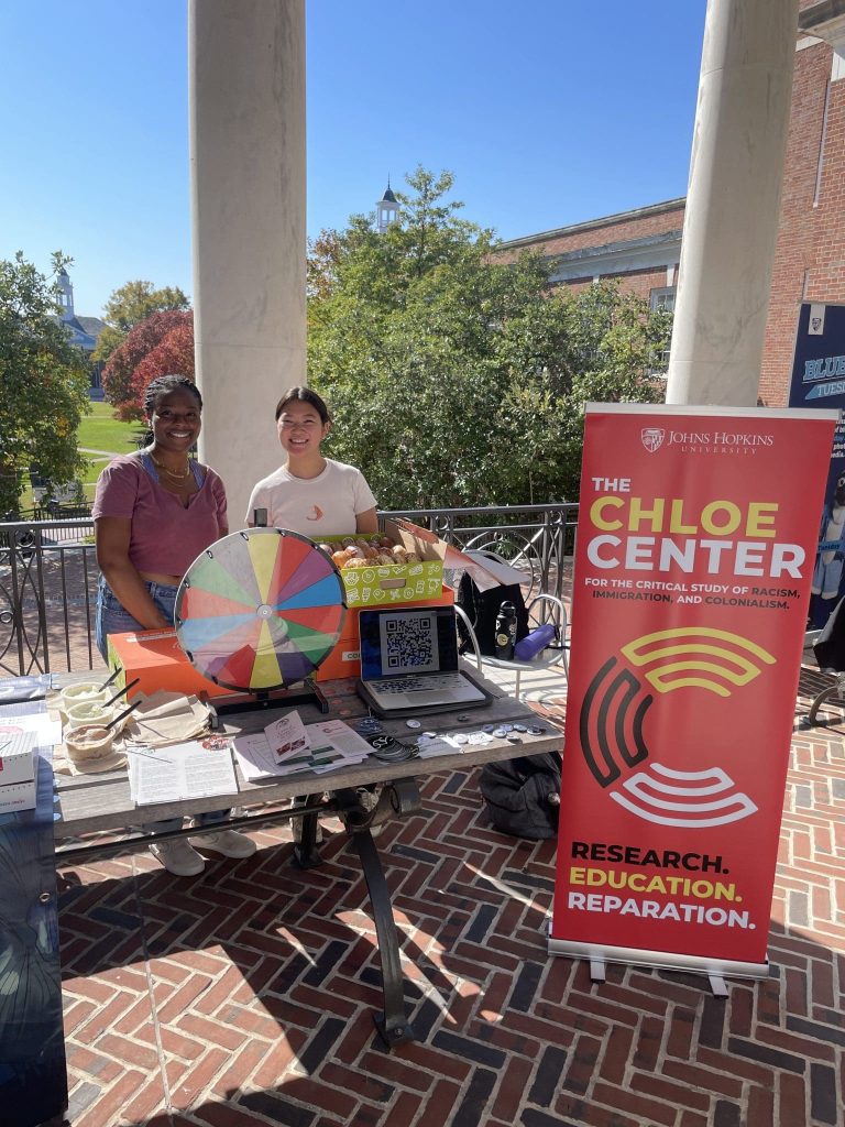 Two students behind a table with a spinning prize wheel, bagels, and papers