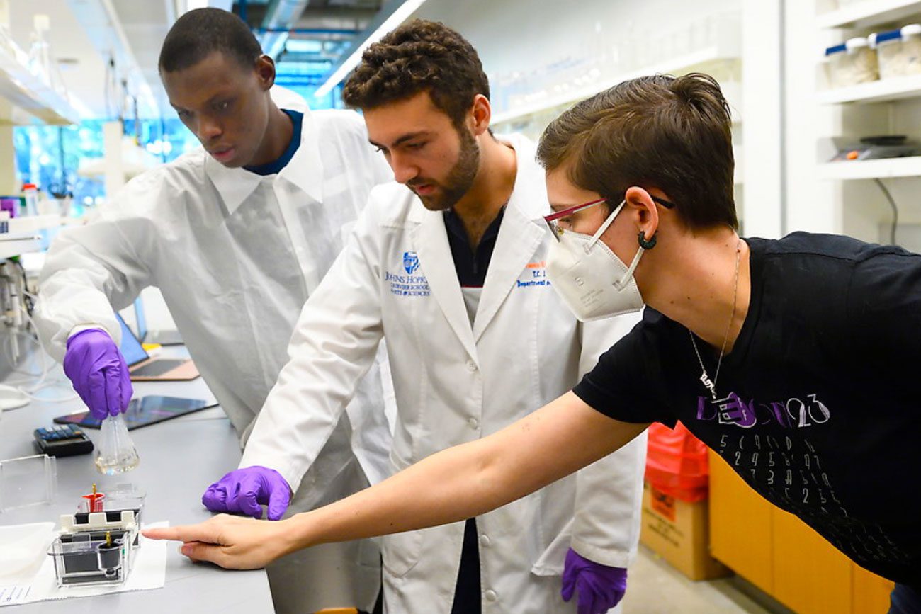 group of three students in lab coats pointing at a device on a counter