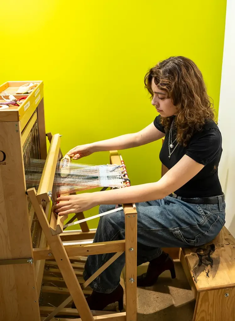 Daniela Rodriguez ’24 seated at a wooden loom, guiding threads through the frame to weave her creative textile design.