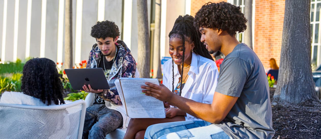 Fours students outdoors on Keyser Quad talking, looking at their laptops on a sunny Spring day.