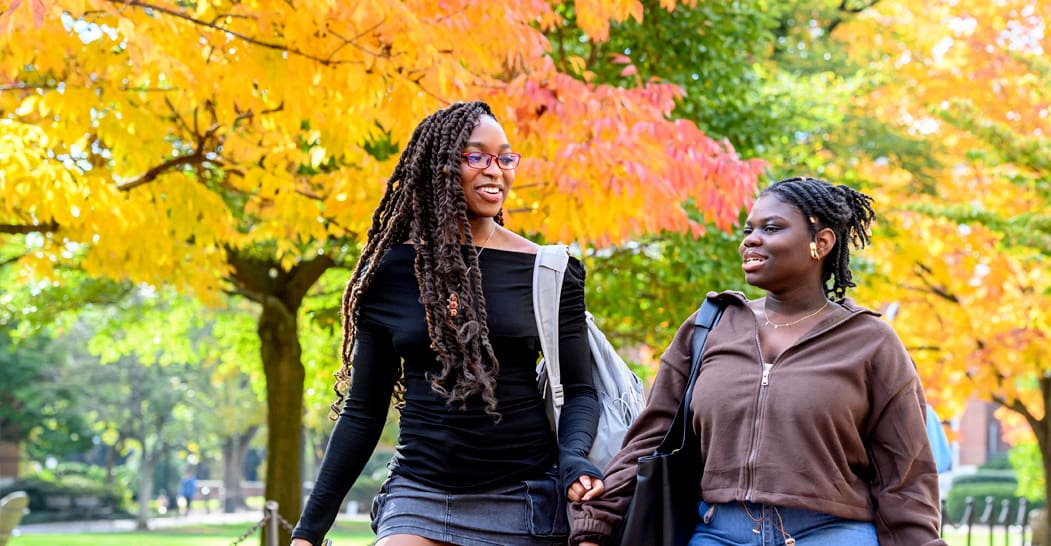 Two students walk outdoors on campus, surrounded by trees adorned in vibrant fall-colored leaves.