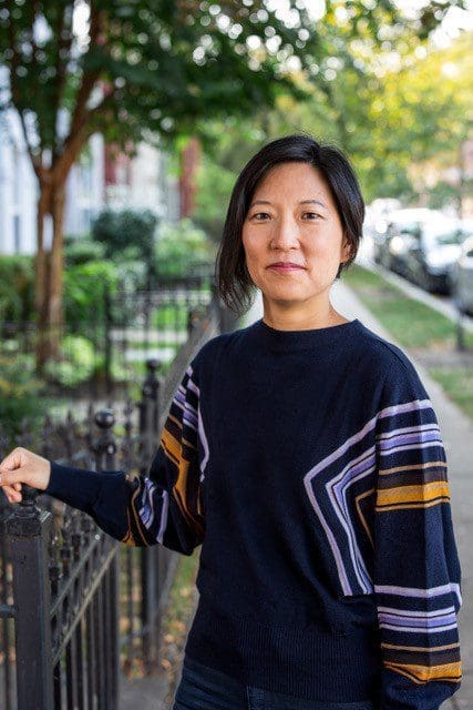 Photo of Clara Han. She is wearing a blue blouse and her hand is resting on an iron fence.