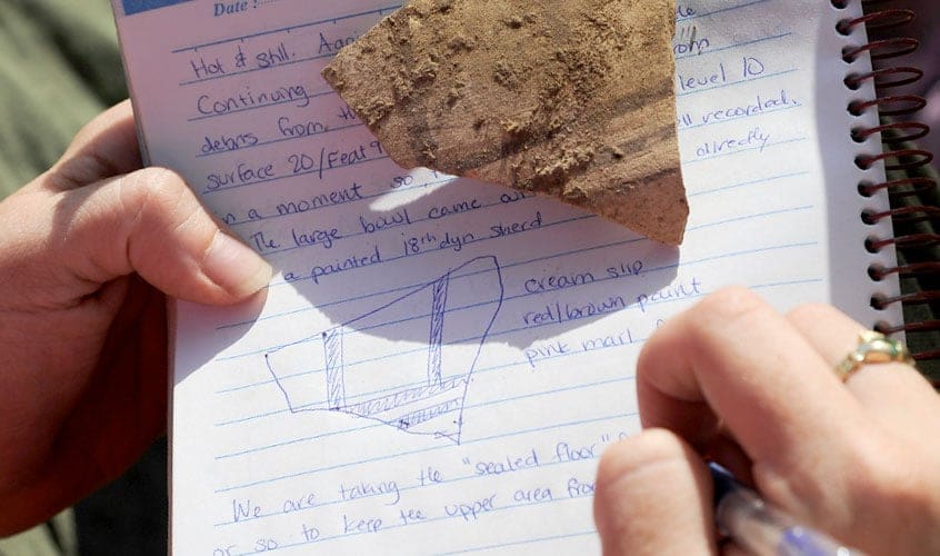 A close-up of student's hands and journal entry with drawing and making notes about a pottery shard discovered on a dig in Luxor, Eqypt.