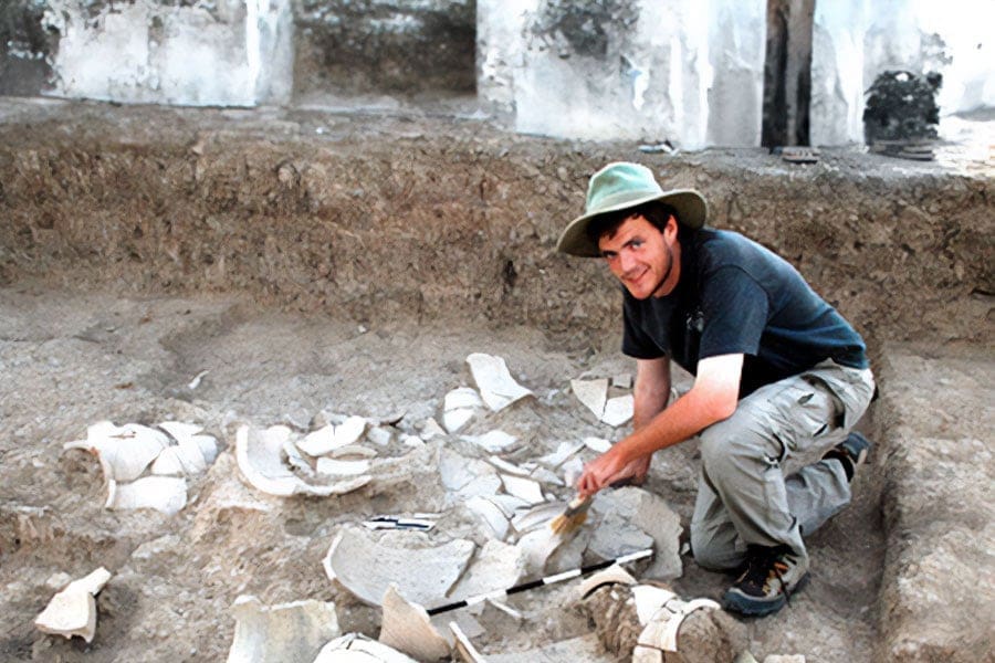 student in hat at dig with pottery shards in dirt