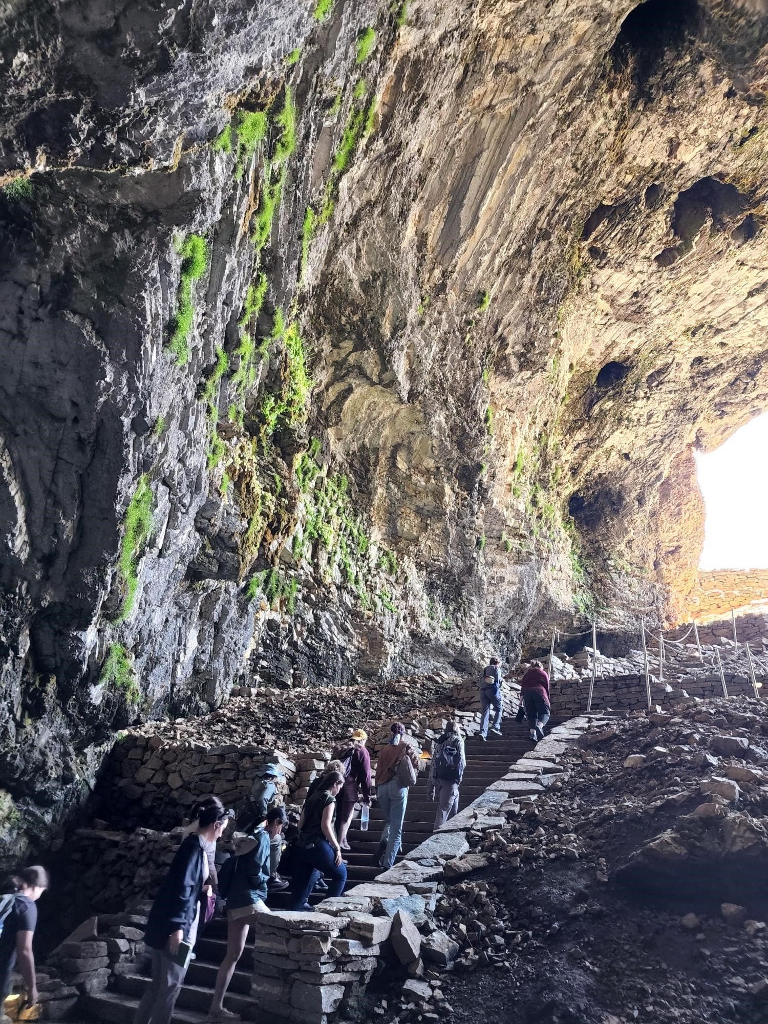 People climbing stone stairs inside a large, moss-covered cave.