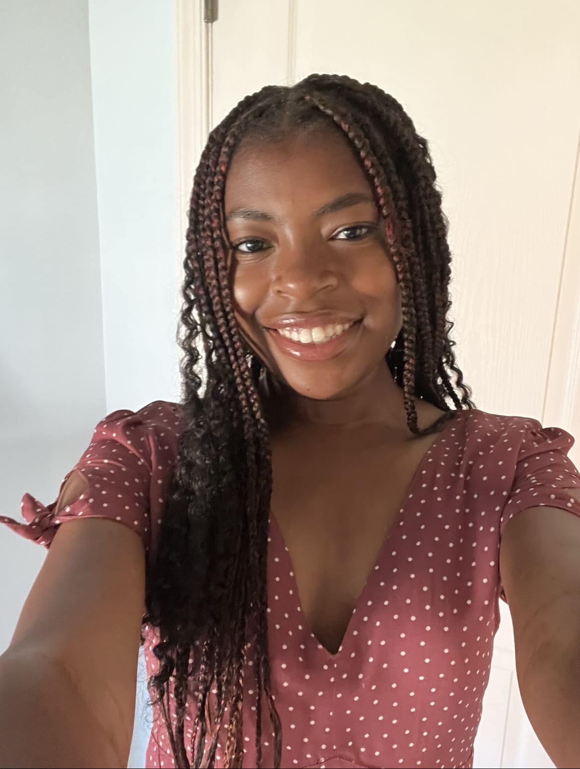 A smiling student holding out the camera to take a selfie. They have long brown braids and are wearing a pink polka dot shirt.