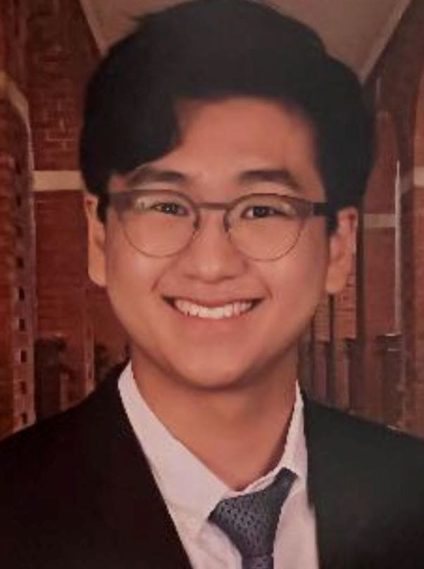 A smiling student with glasses and black hair styled to one side. They're wearing a dress jacket and tie.