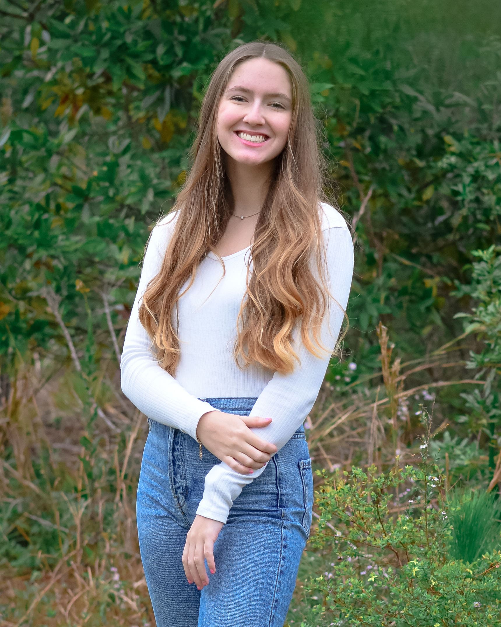 Smiling student takes a photo in front of large, green bushes. They have long brown hair and are wearing a white long sleeve shirt.