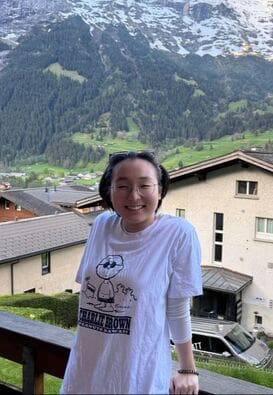 A smiling student with glasses up on forehead, pushing back their hair. They are standing in front of a mountainous scene with a tan house behind them. A charlie brown cartoon is on their shirt.