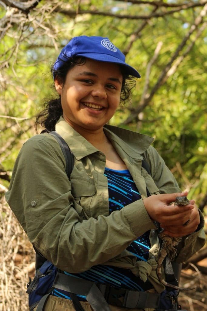 Aarushi stands in the center of the photo with a blue cap and green jacket on. A big smile is on their face as they hold the species they researched. It is small and lizard like, staring at the camera