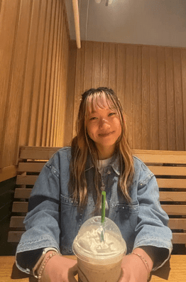 Kaitlyn smiling at the camera in a blue shirt with her hands around a frozen coffee