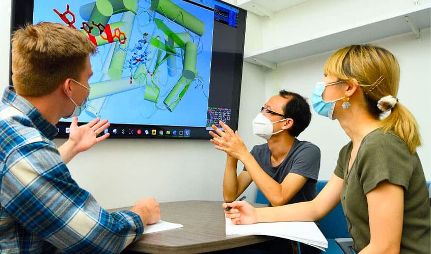 Chemistry professor Xiongyi Huang (center) looking at large monitor with two graduate students.