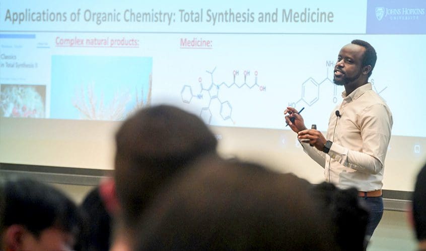 Professor Olivier Nsengiyumva in front of projection screen teaching Organic Chemistry II class.