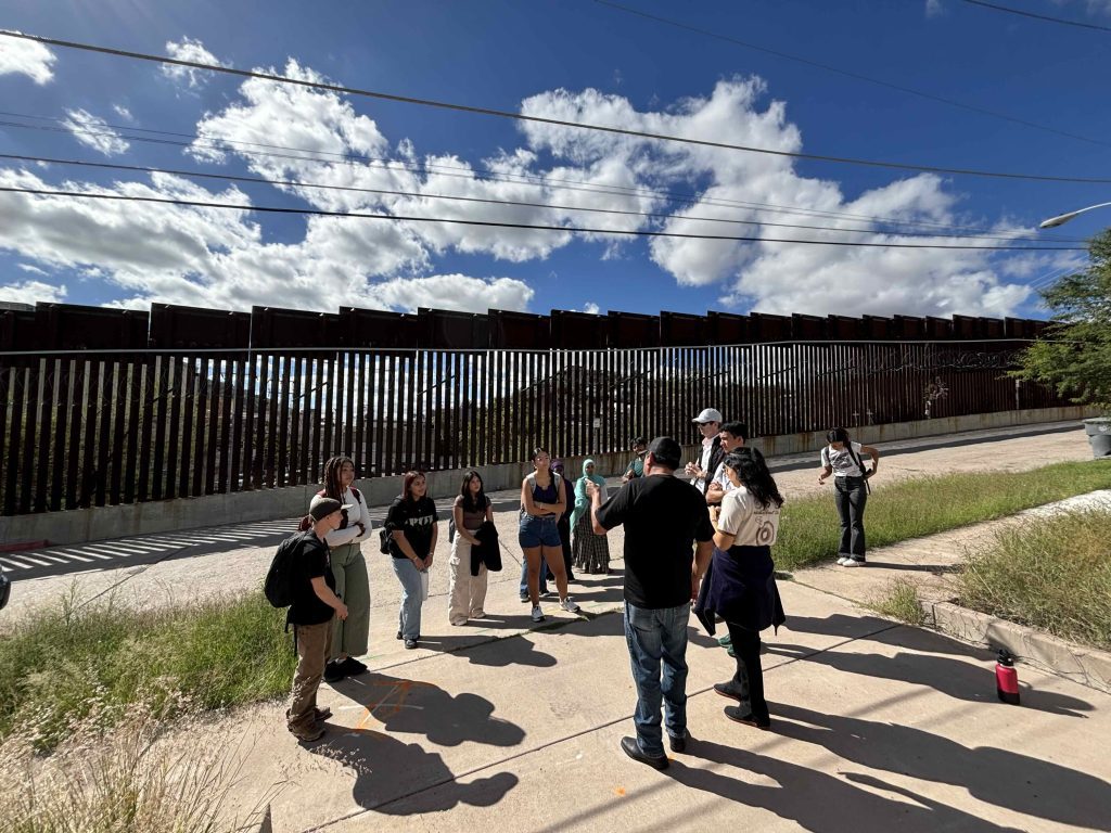 Students listening to speaker in front of US-Mexico border wall, with blue sky and clouds above