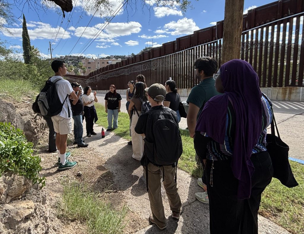 Students listen to speaker in front of US-Mexico border fence with blue sky and clouds above