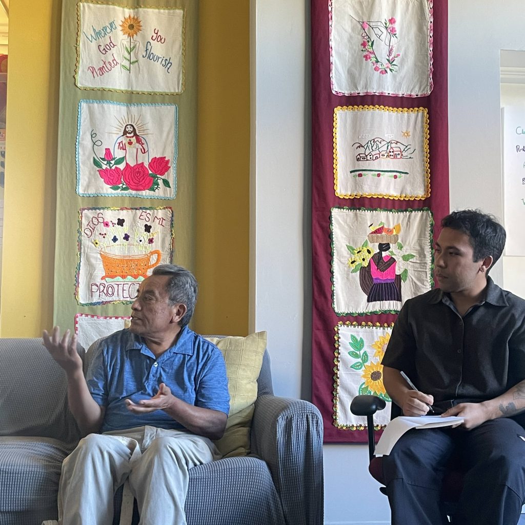 Two men seated in front of decorated wall with fabric mantas with colorful embroidered designs