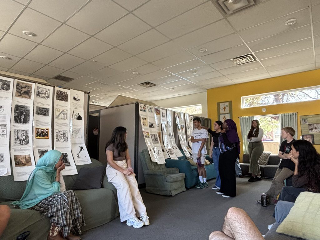 Students in room with couches, standing before posters about immigration history