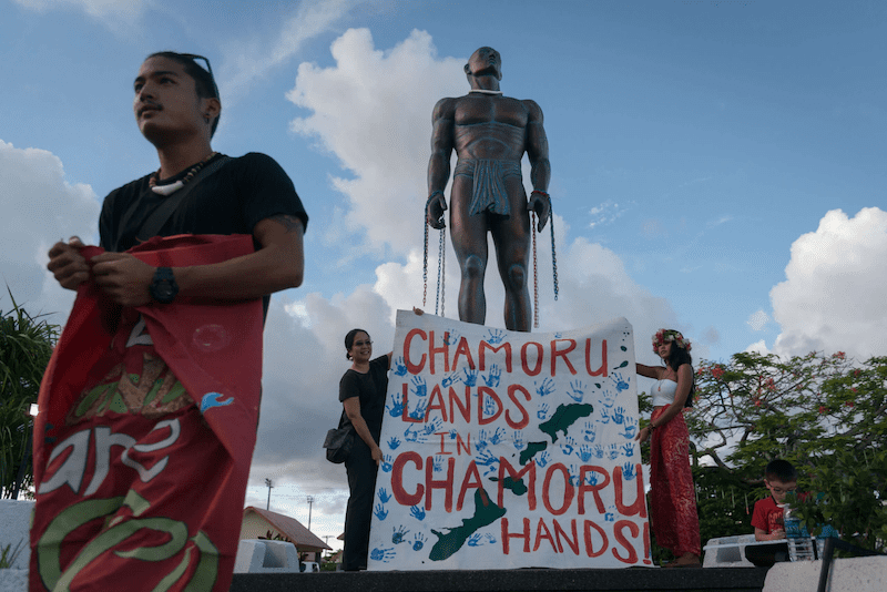 Protesters in front of statue with banner that reads Chamoru Lands in Chamoru Hands