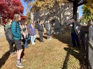 Students standing in a semicircle in a cemetery with fall foliage while a man talks