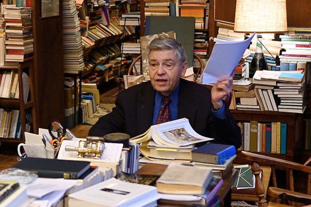 Richard Macksey seated at a desk in a library piled with books