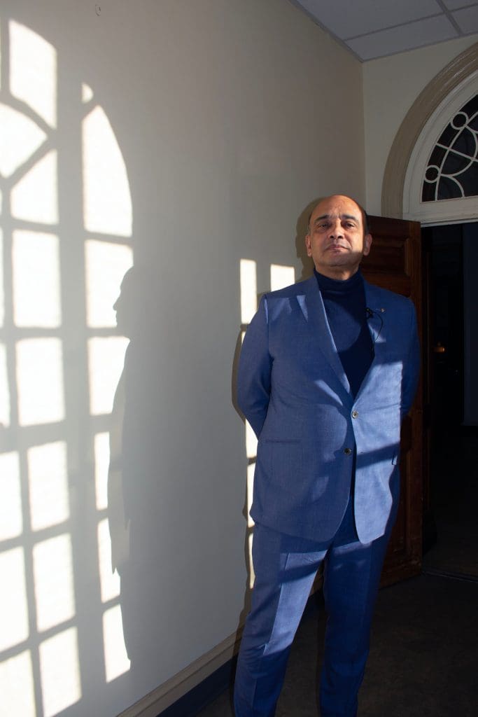 Kwame Anthony Appiah (man in blue suit) standing in afternoon sunlight with his profile framed in shadow and light against the wall.