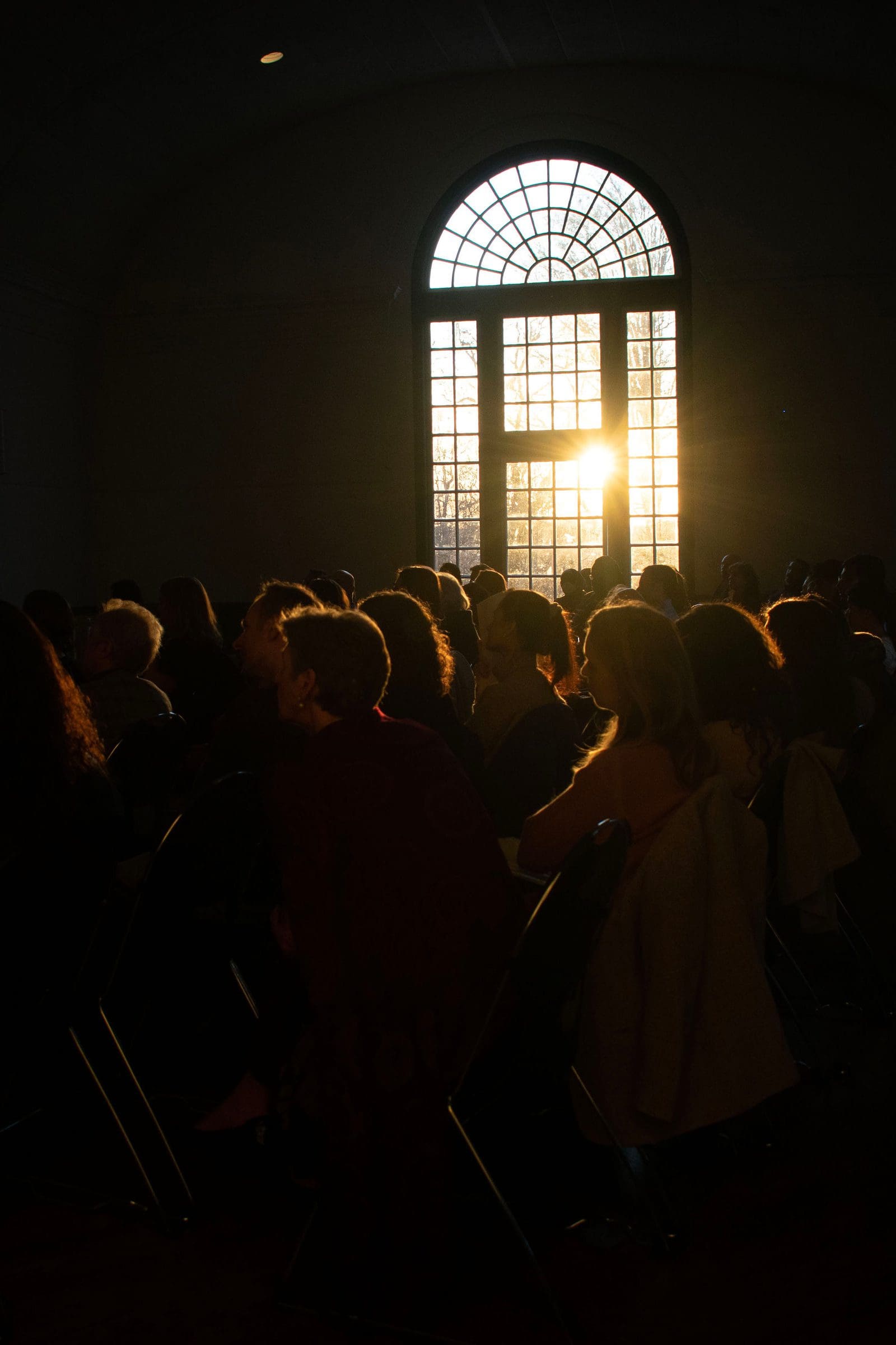 crowd of people in a dark room with a large window letting in sunshine