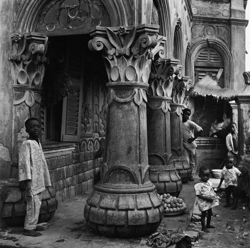 Photography in black and white of young child in Lagos, Nigeria in 1952 standing beside a highly ornamental Afro-Brazilian column (with other local children in the background).