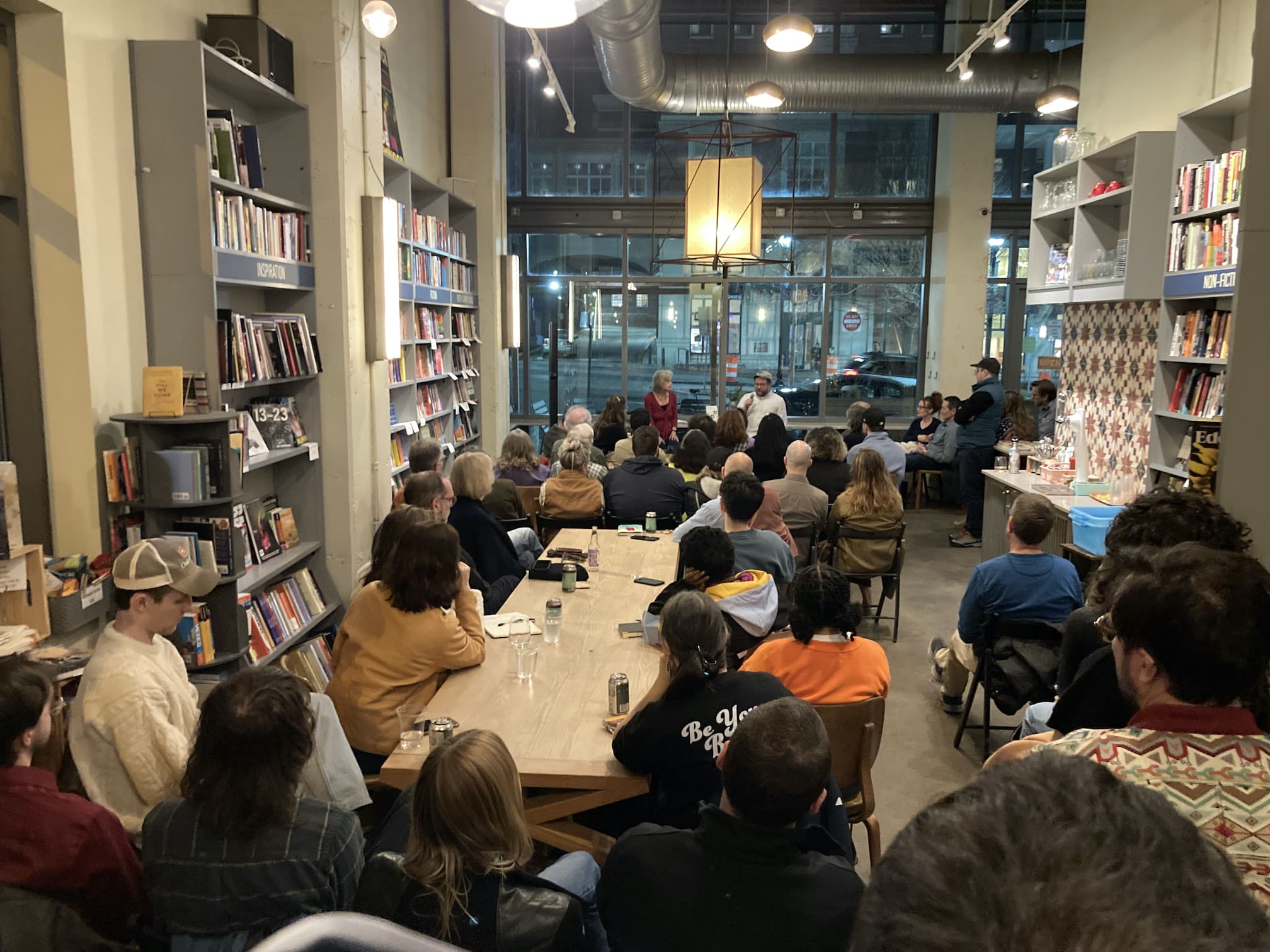 Crowd viewed from the back of a long, packed bookshop, with two speakers (left, an older woman; right, a capped man speaking into a microphone) in the background of Bird in Hand cafe.