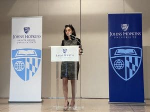Speaker Merve Emre presents at a podium in front of two JHU KSAS banners at the March 2024 Macksey Lecture in the Glass Pavilion.