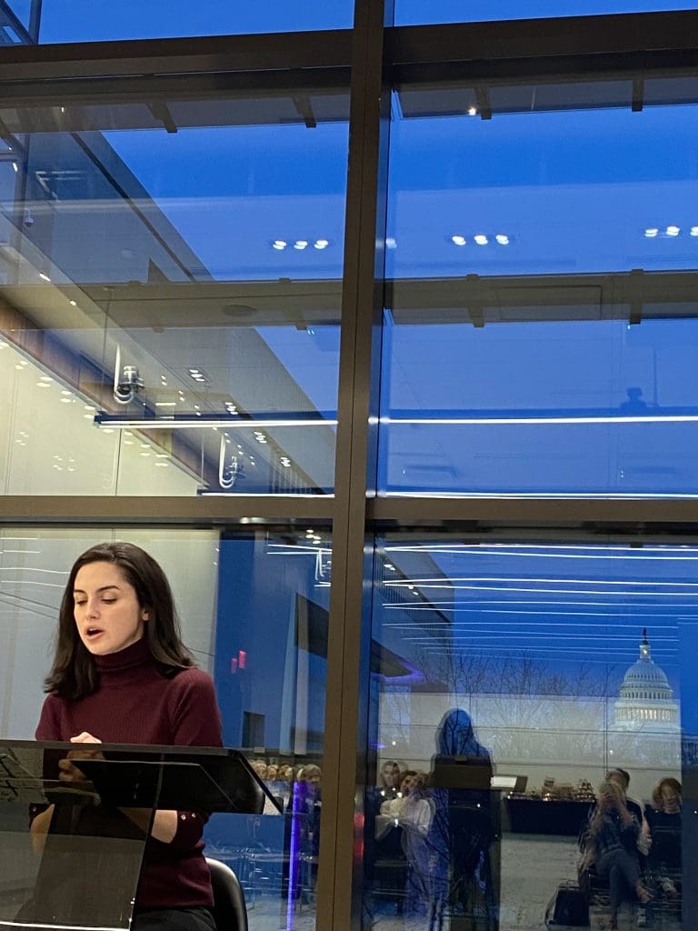 Merve Emre sits at a podium to read her talk on Woolf and Bourdieu, sitting in the JHU Bloomberg Center with the Capitol Building and DC skyline at evening behind her.