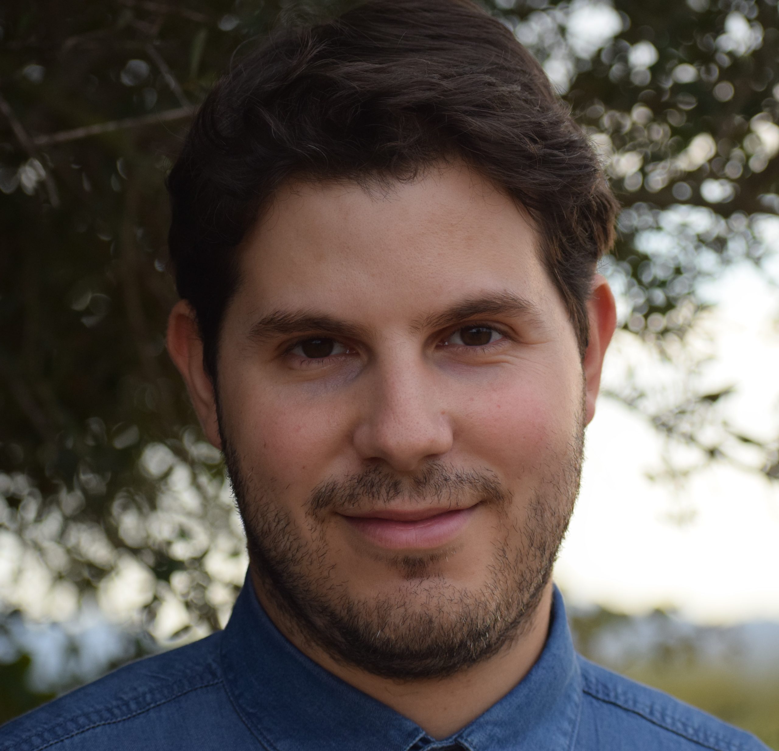 Giacomo Loi wearing a denim button up shirt with trees behind him