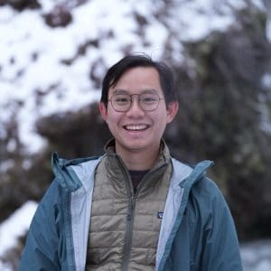 Arthur with blurred trees in the background wearing a blue coat over a  light green puffer vest and eye glasses
