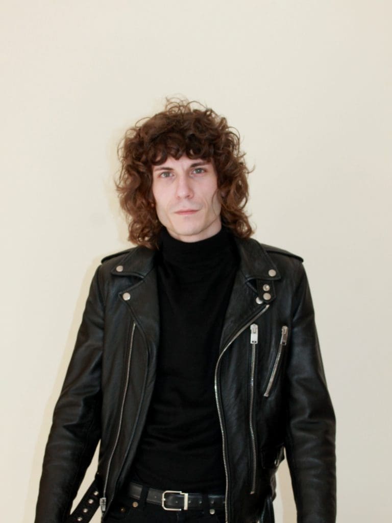 Gianluca wearing leather jacket and black shirt and curly shoulder length hair