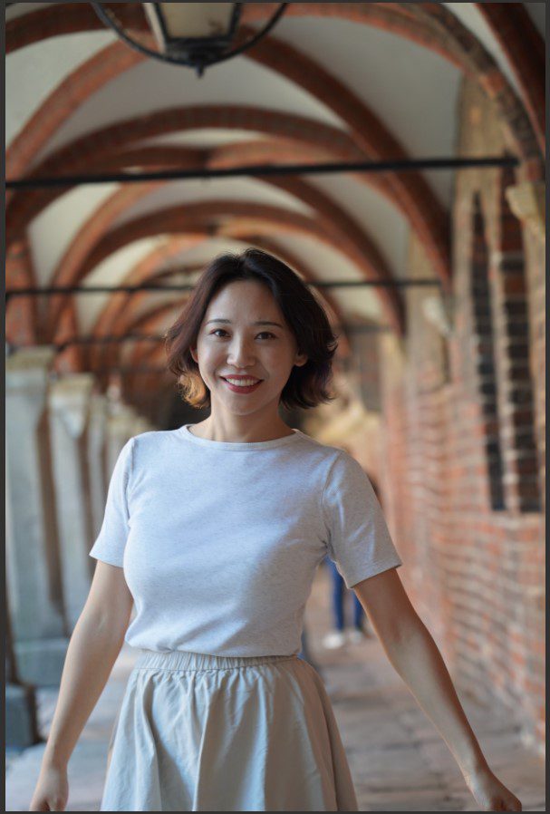 Portrait of Shengshuang Wang standing in a brick arched walkway, wearing a light-colored top and skirt.
