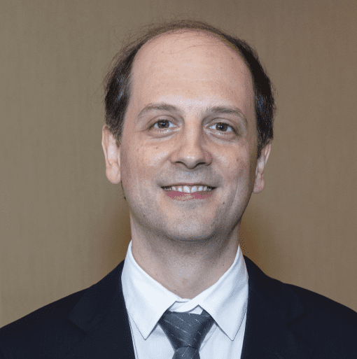 Dr. Rutishauser is smiling facing the camera wearing a black business jacket with white collared shirt and tie.
