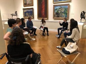 students in a circle of chairs in the middle of a room in an art museum