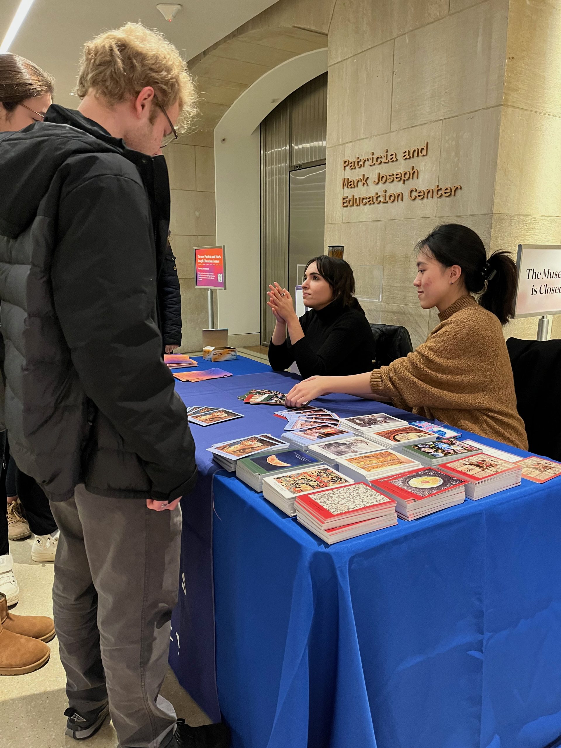 Two students sit at an event sign-in table and face a standing student. Postcards appear on the table.