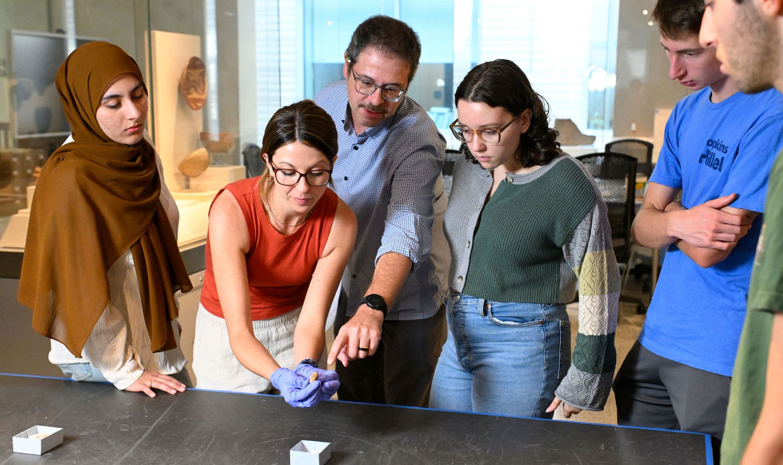 Faculty (Jacob Lauinger) in the center pointing towards an artifact that a person with a red shirt and gloves is holding. Students surround them.