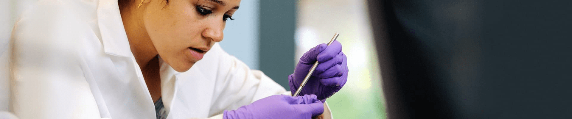 a female student wearing a white lab coat and purple gloves holds a tool