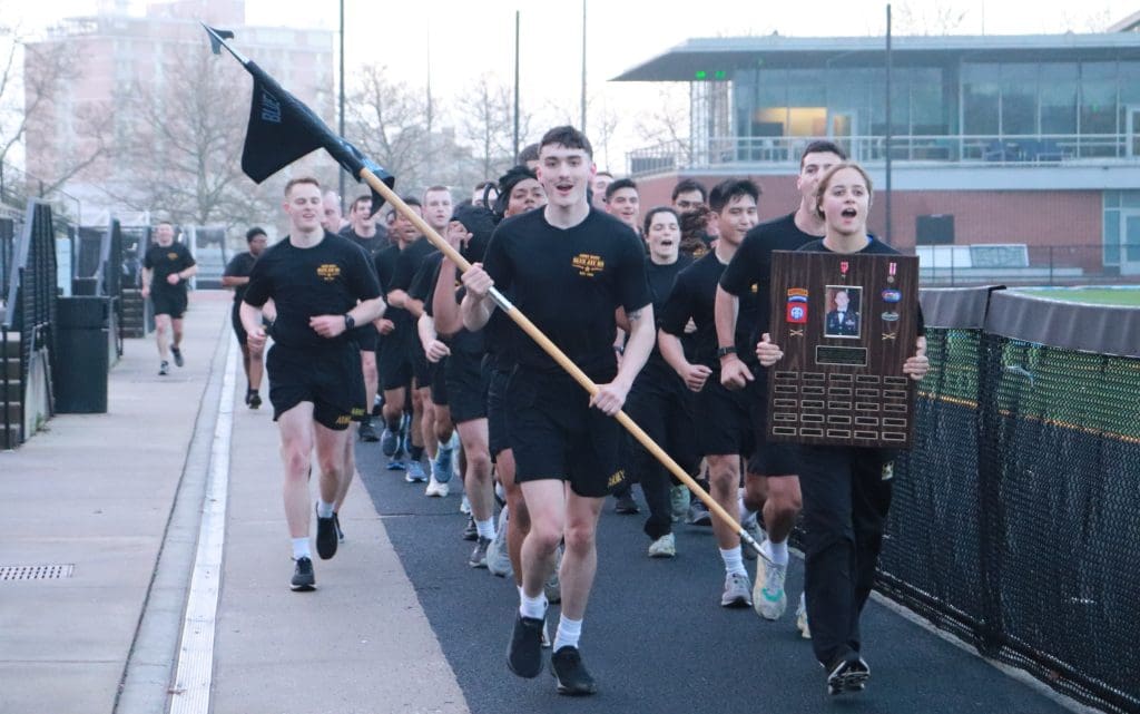 group of cadets running on the track with one in the front carrying a flag and another carrying a plaque with CPT Grassbaugh's picture