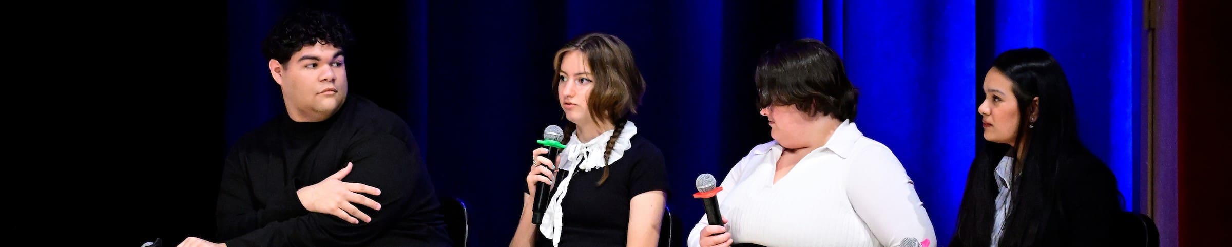 A panel of four students sits on stage in Shriver Hall Auditorium. One is speaking into a microphone and the other three are listening.