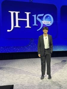 Sareen Muthyala stands before a screen with the JH150 logo