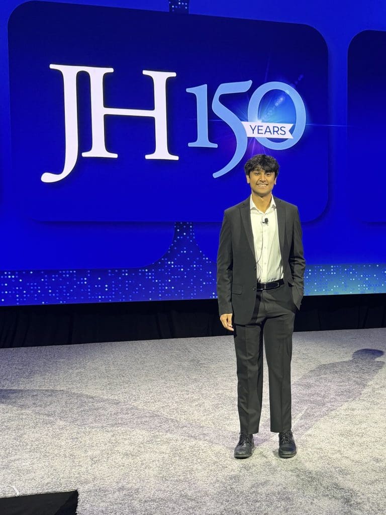 Sareen Muthyala stands before a screen with the JH150 logo