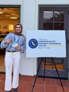 Arusa Malik stands next to a sign announcing the 24th annual Undergraduate Research Conference on the European Union, April 9-10, 2026.