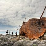 a close up photo of a washed up rusted ship on rocks with six figures in the background