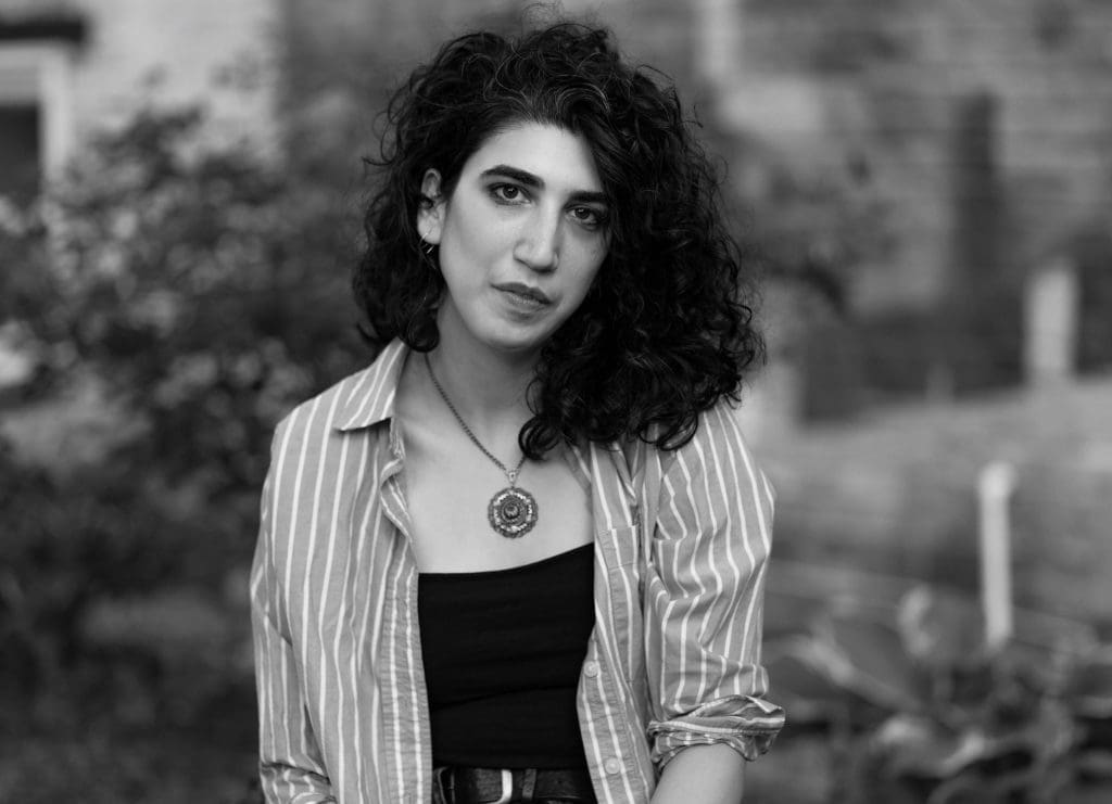 Black and white bust portrait of a woman with dark curly hair, wearing an opened button down striped shirt with a black shirt underneath. She is wearing a large necklace and is standing in nature with plants behind here, out of focus.