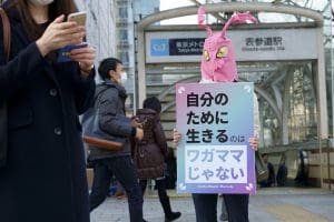 a person wearing a pink, rabbit-like mask with a sign in front of a busy Japanese train station