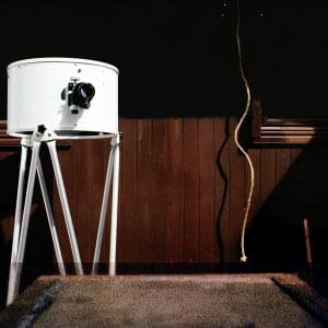 Color image background top half is black bottom half contains brown wood wall paneling and beige carpet below. In the foreground there is a large white camera with thripod legs from the Ukiah Latitude Observatory in Ukiah, California on the left of the image and a tan rope hanging down the right side of the image with a shadow next to the bottom of a a wooden window frame.