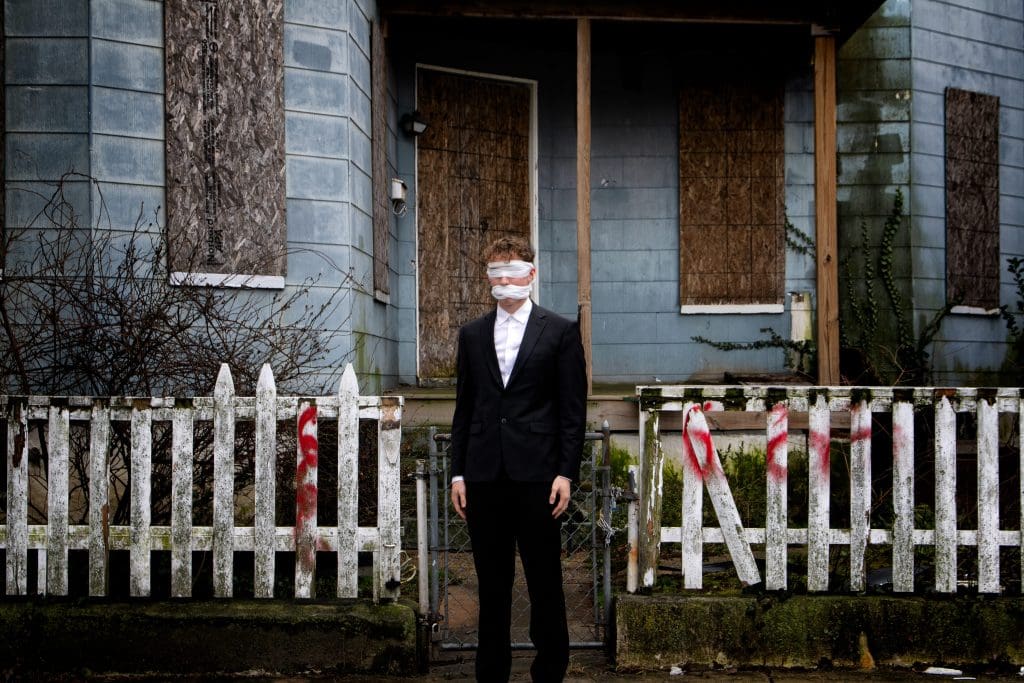 a blindfolded man wearing a suit in front of a boarded up house