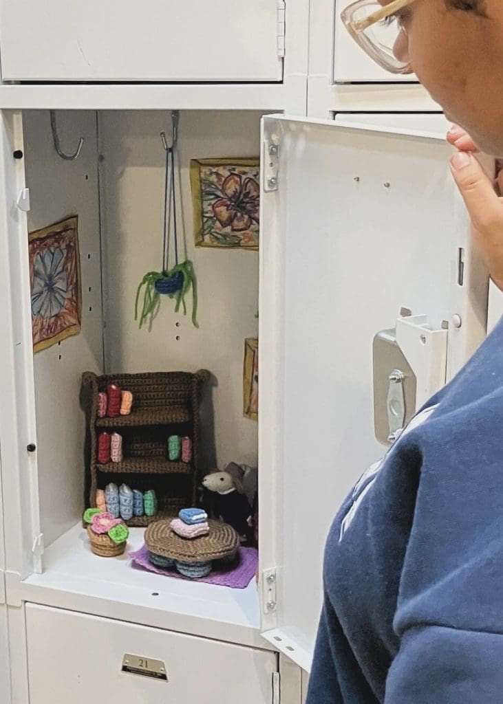 student showing their artwork of a crochet mouse, bookshelf, table, rug, and plants installed in a small metal locker